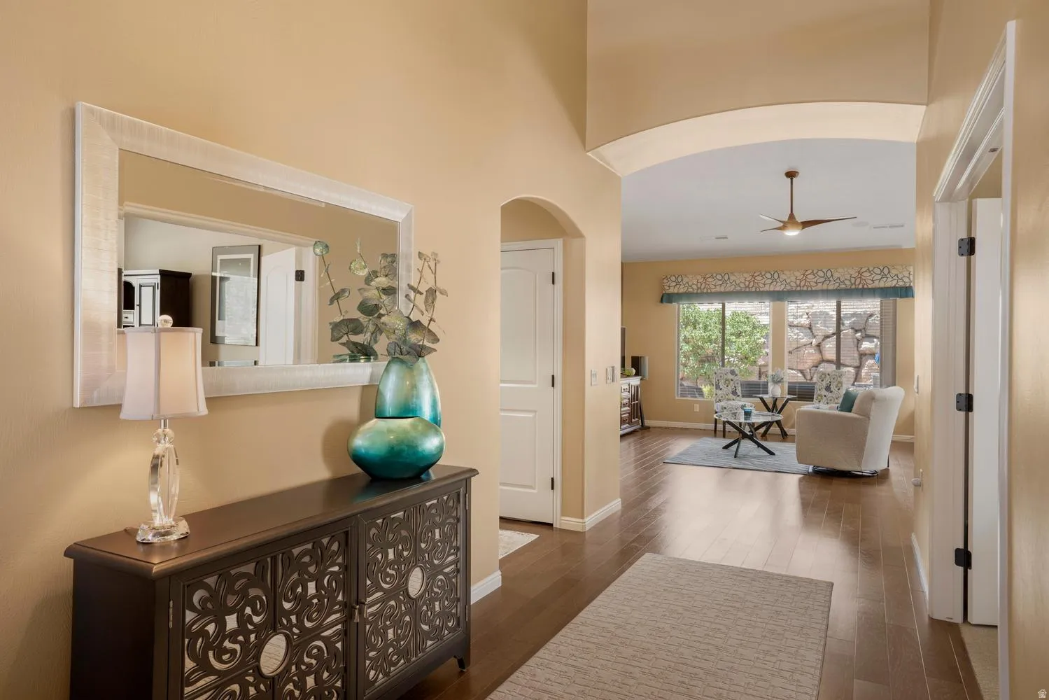 Arched entryway leading to a spacious living area featuring wood-finish flooring, a ceiling fan, and large windows with custom window treatments