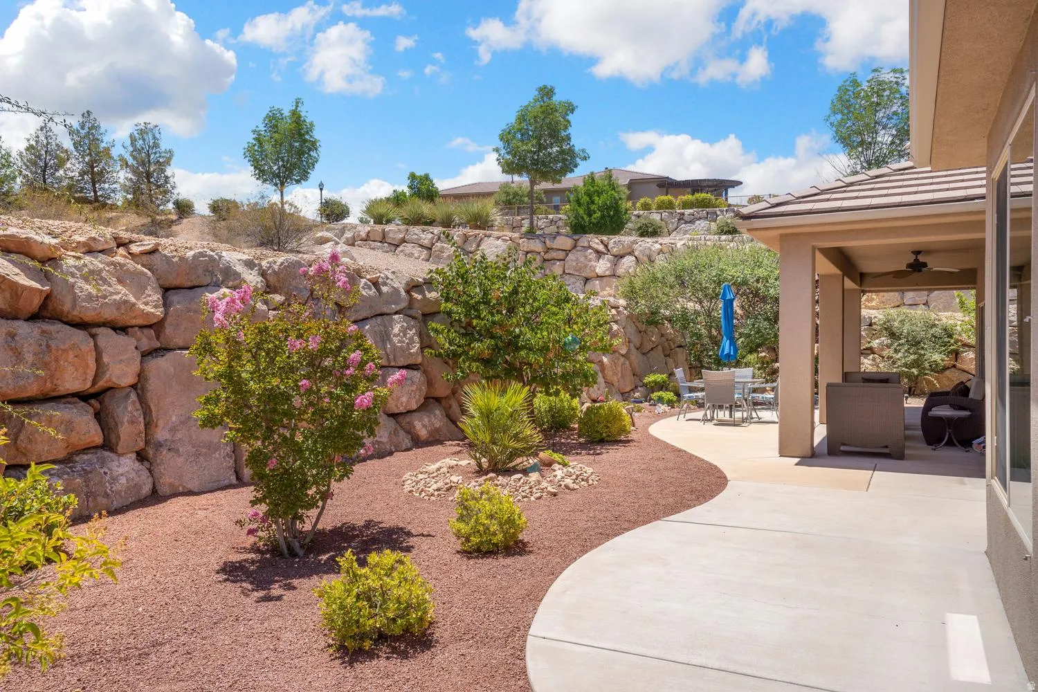 Expansive outdoor patio with a covered section, concrete hardscaping, rock retaining walls, and mature landscaping