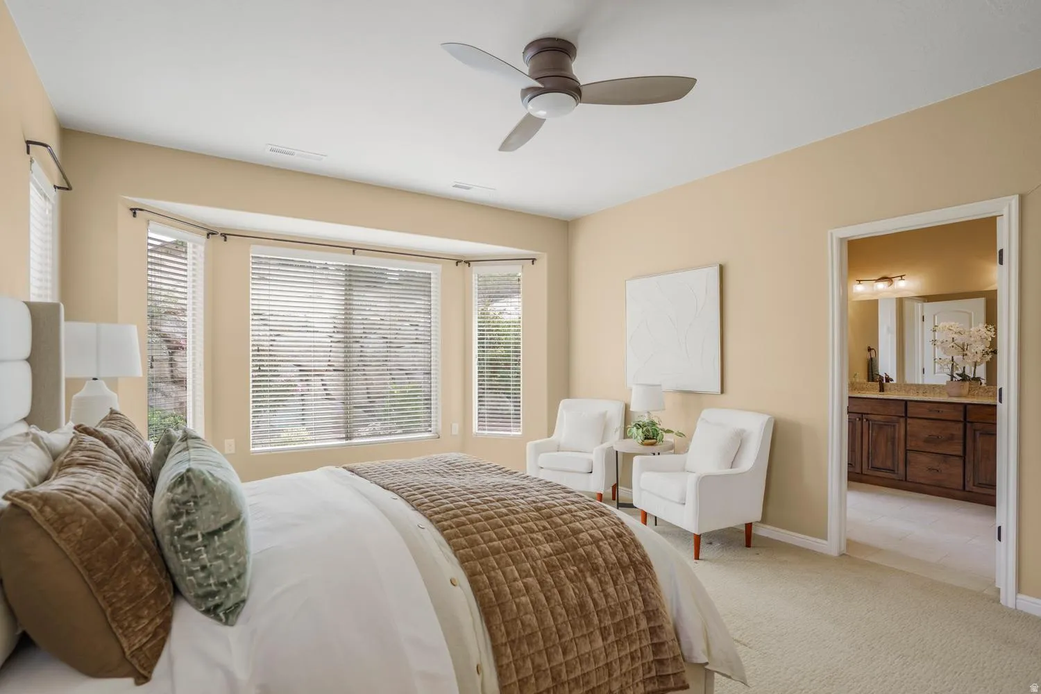 Recessed bay window, ceiling fan, carpet flooring, a doorway leading to a room with a wood-finish vanity, and a mirror