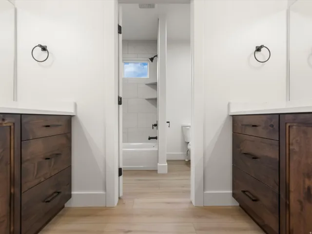 Bathroom featuring dual wood-finish vanities with light countertops, light wood-finish flooring, and a white bathtub with subway tile surround