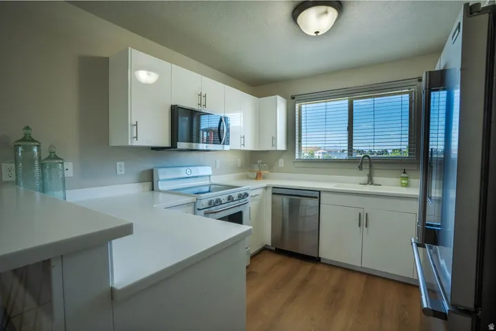 Modern kitchen featuring white cabinetry, light-toned countertops, stainless steel appliances, wood-finish flooring, and a large window with blinds