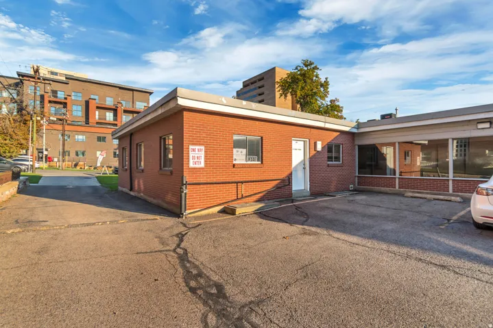 Brick exterior building with multiple windows, a white entry door, and an attached parking area
