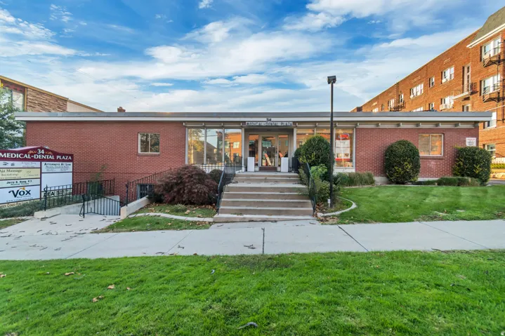Brick facade building featuring a prominent glass storefront entrance, concrete steps with handrails, a secondary accessible ramp, and landscaped shrubbery