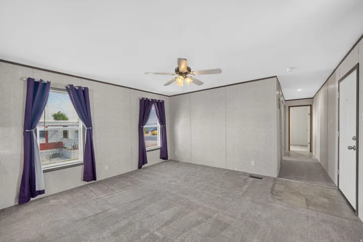 Spacious room featuring gray carpeting, a ceiling fan with integrated lighting, two windows with blinds, and a hallway entrance