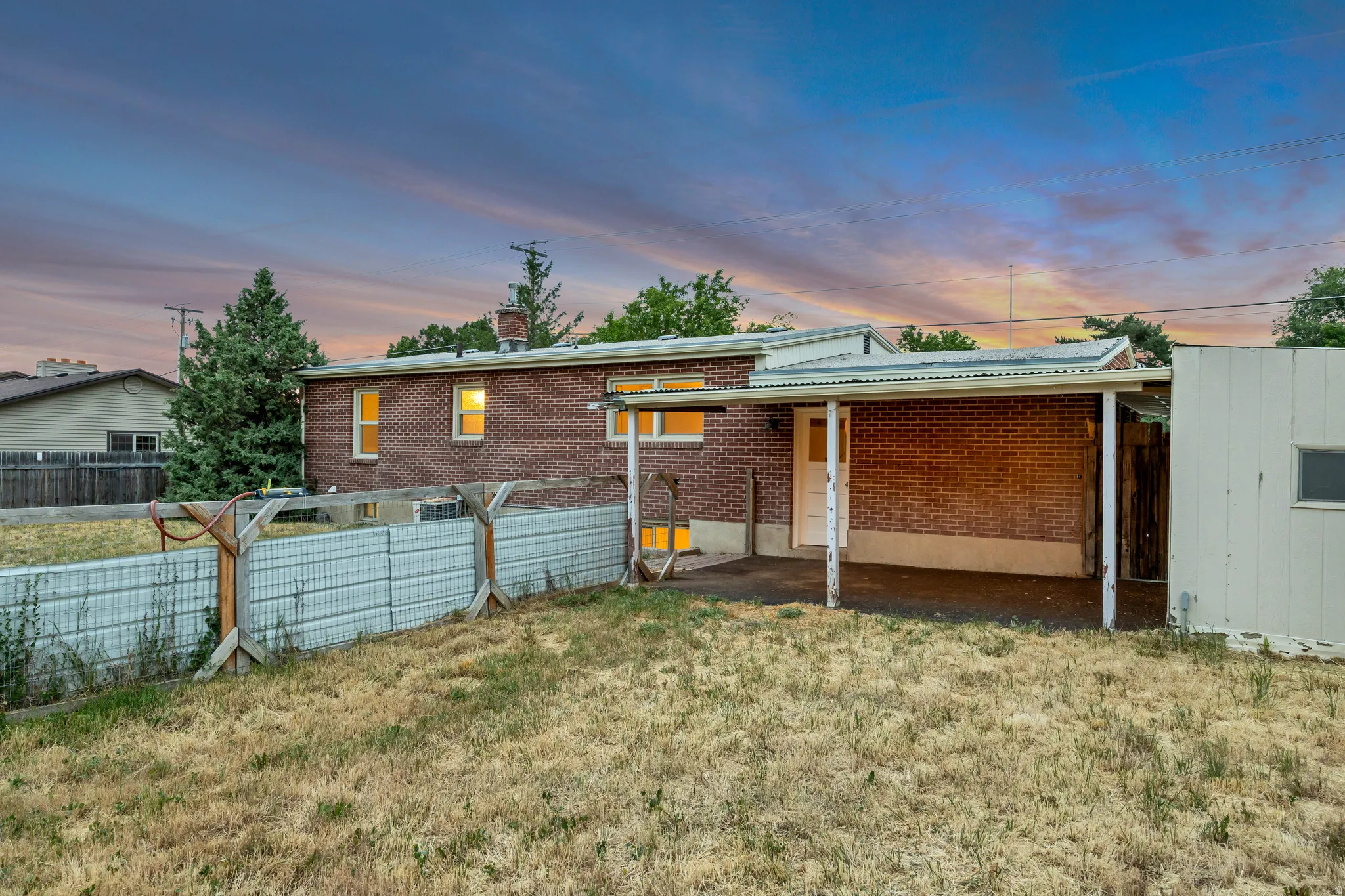 Brick exterior with a covered patio area, mature trees, and a metal privacy fence