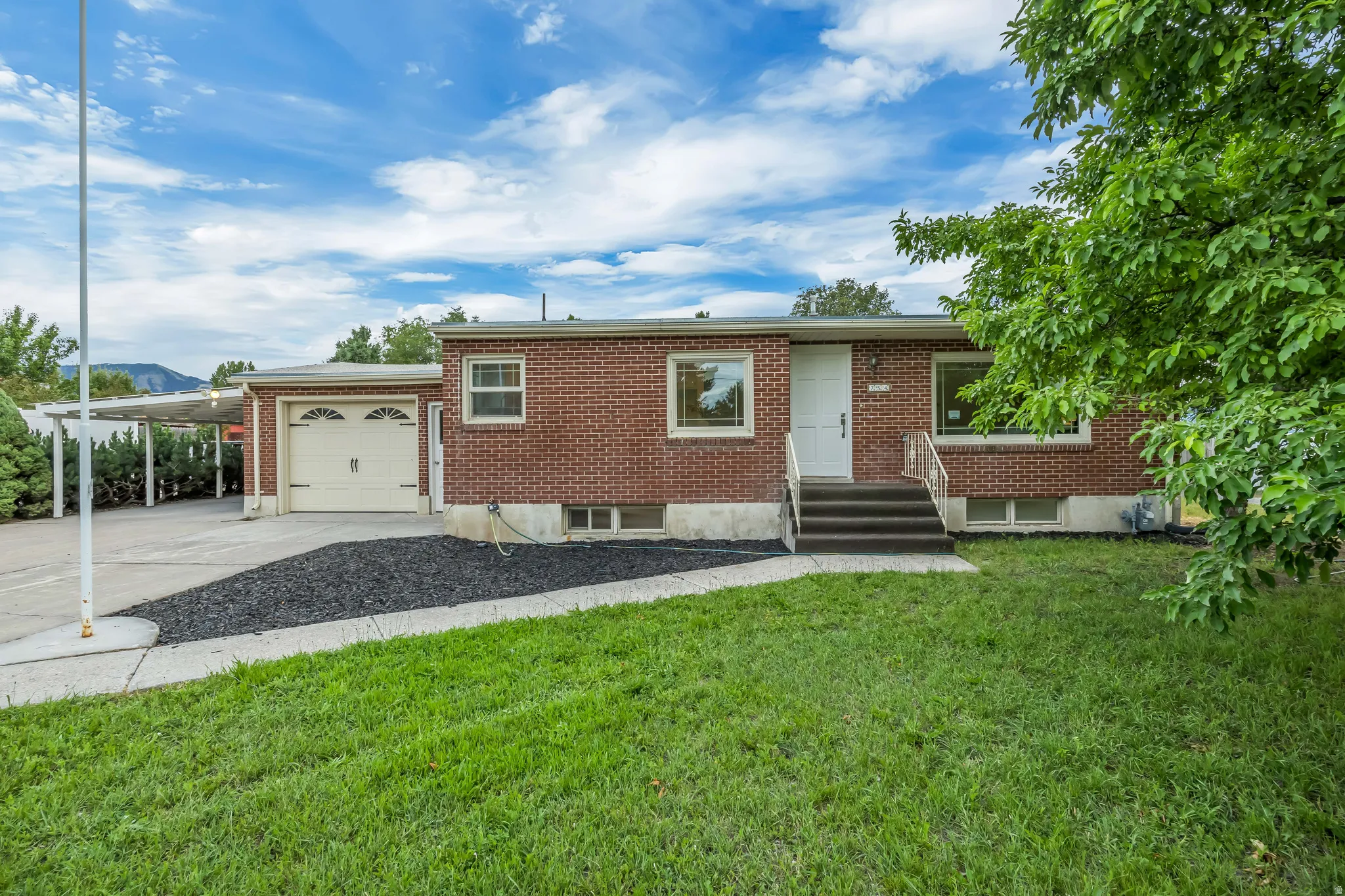 Brick-clad exterior featuring a single-car garage with a panel door, an attached carport, and a front entry with steps and a white railing