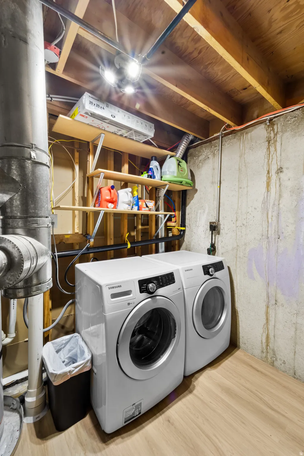 Dedicated laundry area featuring a front-loading washer and dryer, wood-finish flooring, exposed ceiling joists, and concrete walls