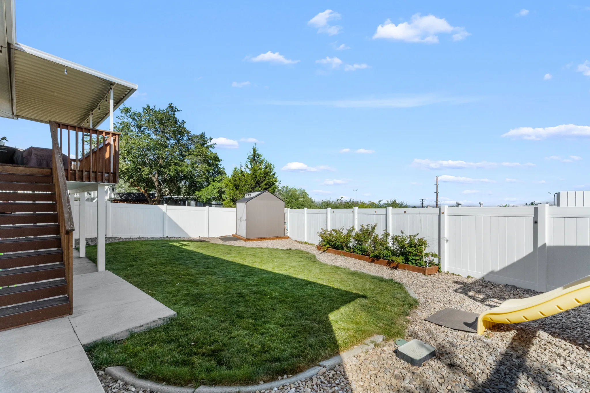 Private backyard featuring a manicured lawn, white vinyl fencing, a gravel landscaping area, and a covered patio structure with wood stairs