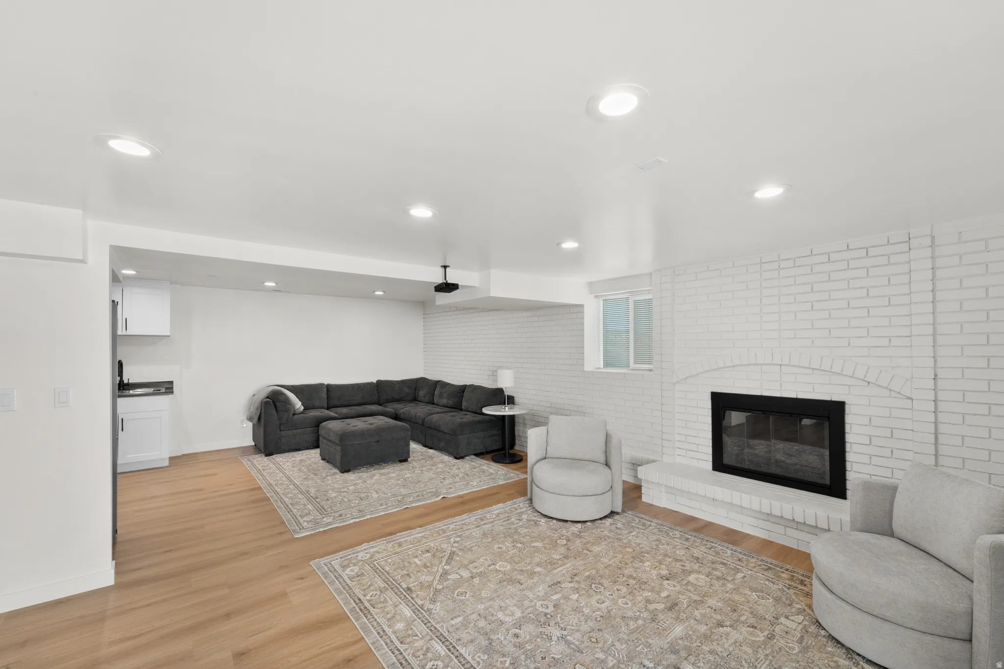 Spacious living area featuring light wood-finish flooring, recessed lighting, and a white-painted brick fireplace with a black insert