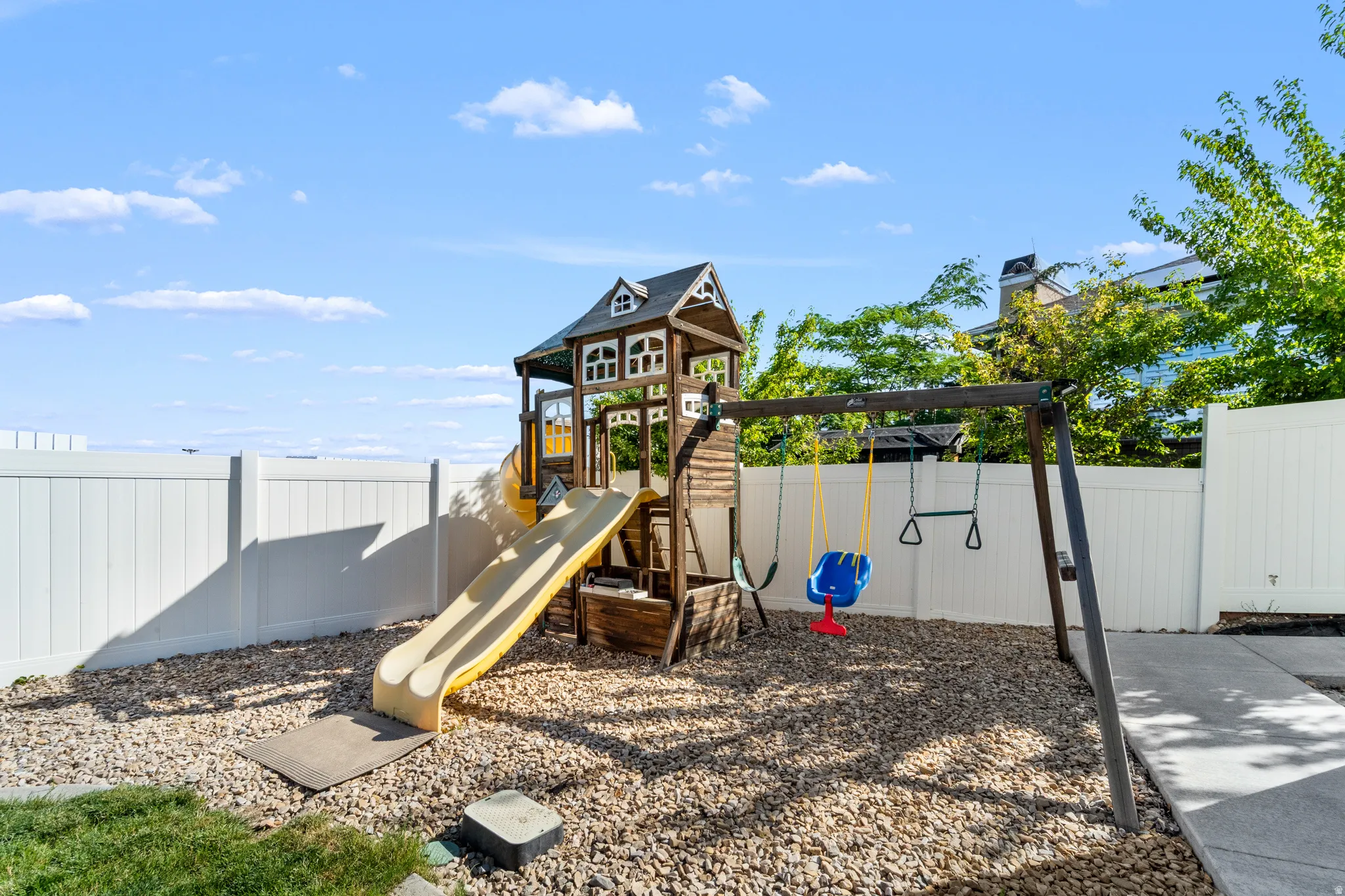 Fenced backyard featuring a wooden playset with slide and swing set, gravel ground cover, and concrete pathway