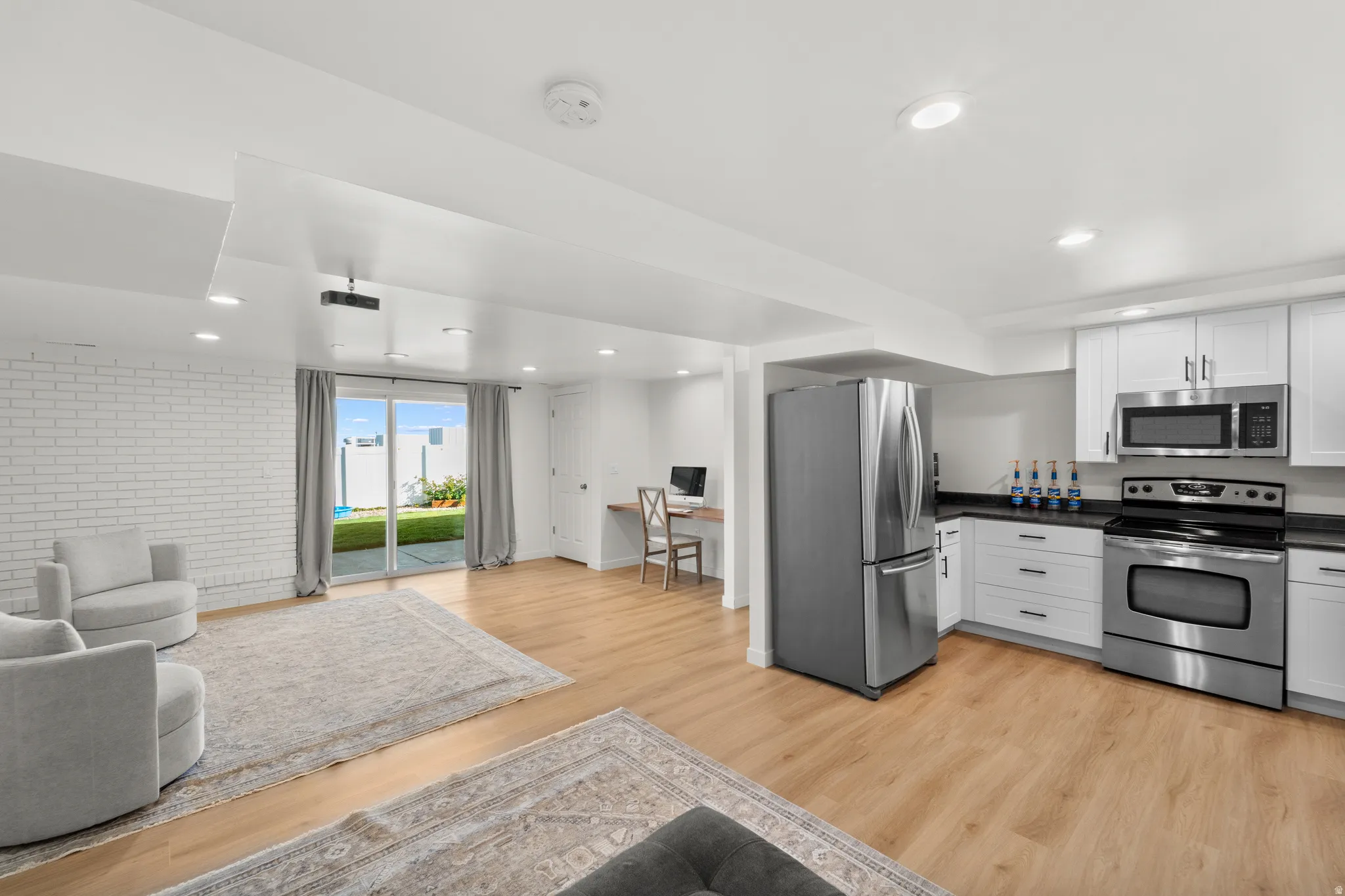 Open-concept living area featuring wood-finish flooring, a white brick accent wall, and a full kitchen with white cabinetry and stainless steel appliances