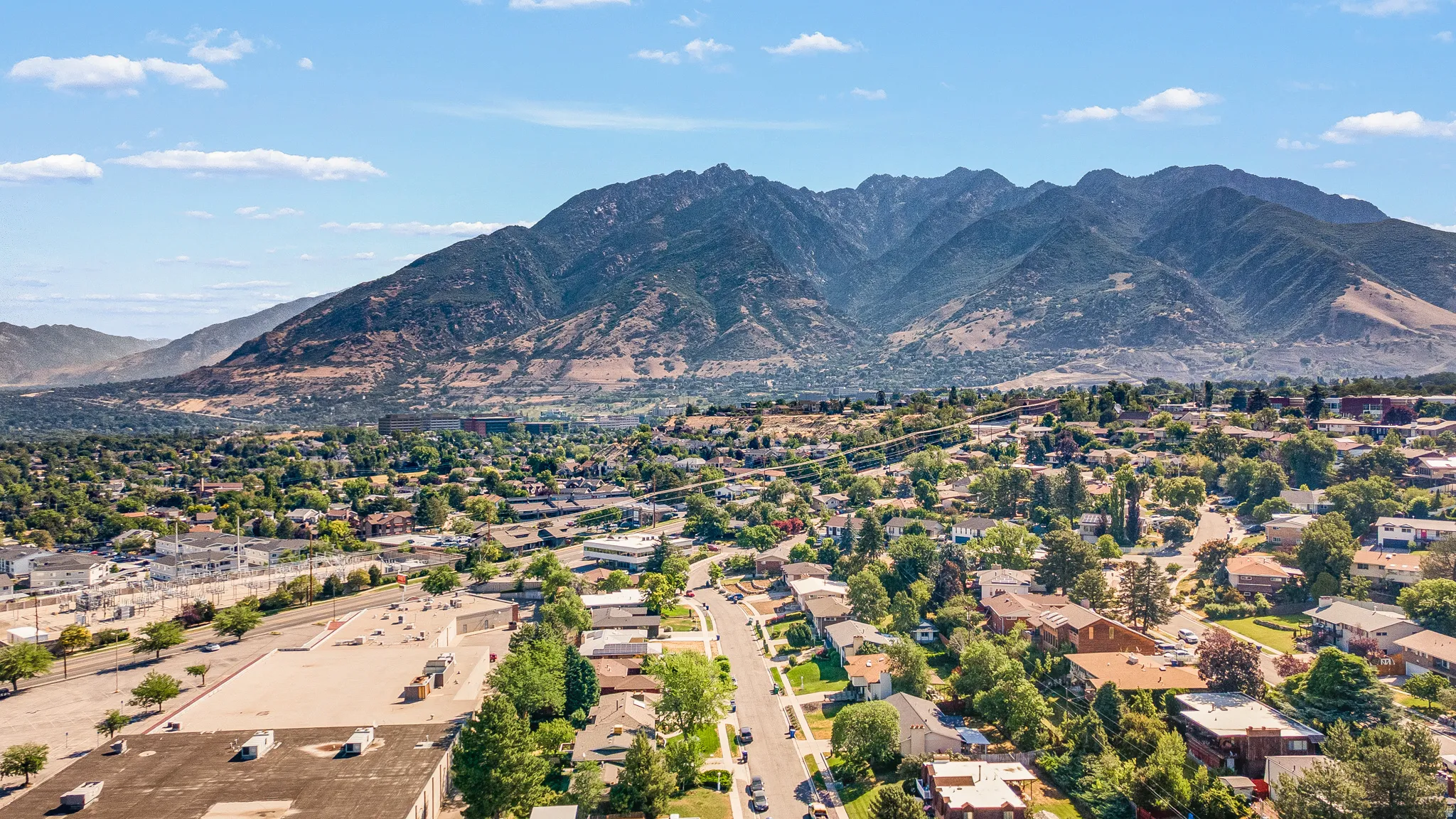 Expansive mountain backdrop framing a residential area with varied rooflines and mature trees, featuring street access and surrounding commercial structures