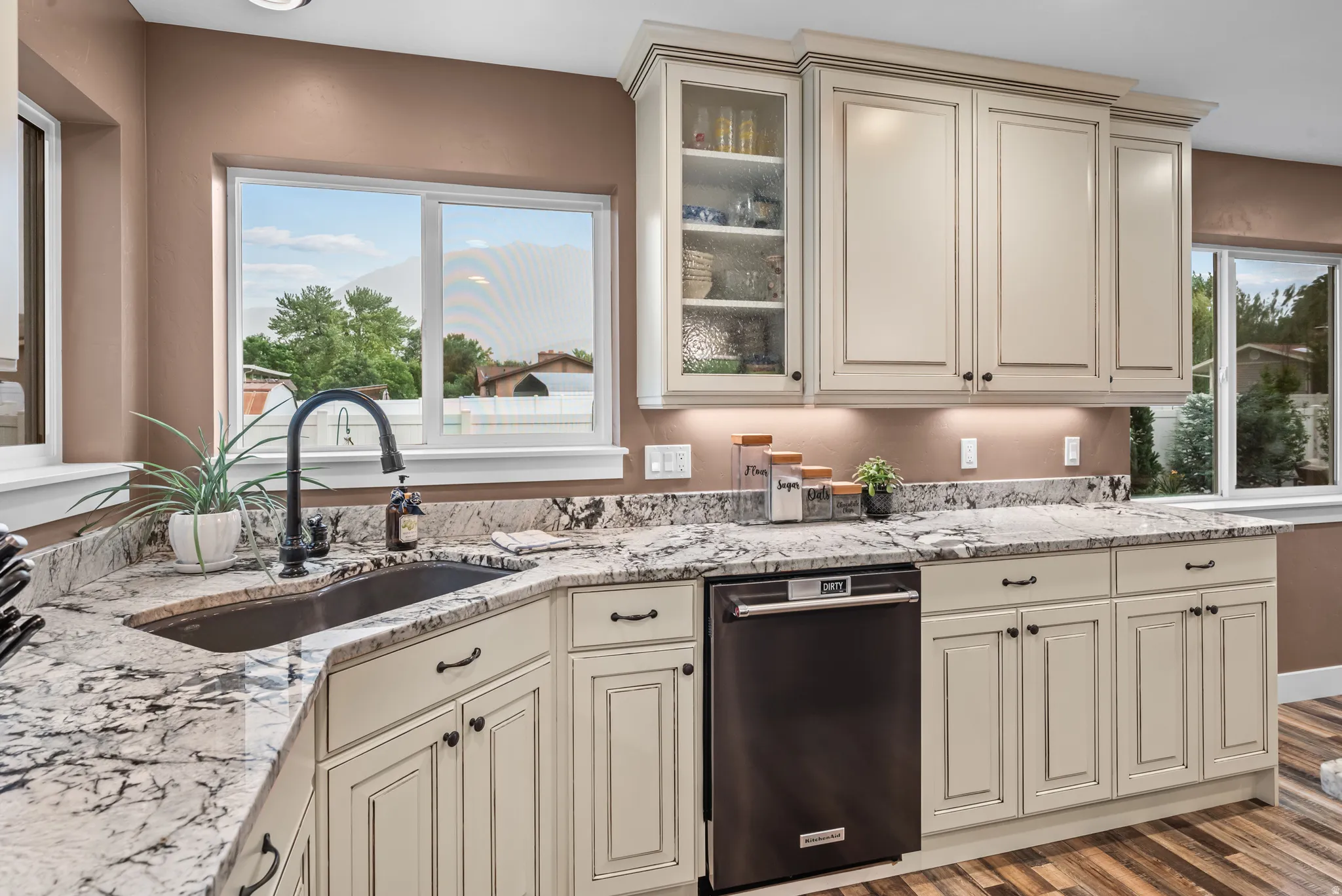 Kitchen featuring stone countertops, undermount sink with a gooseneck faucet, light-colored cabinetry with display glass, under-cabinet lighting, and a built-in dishwasher