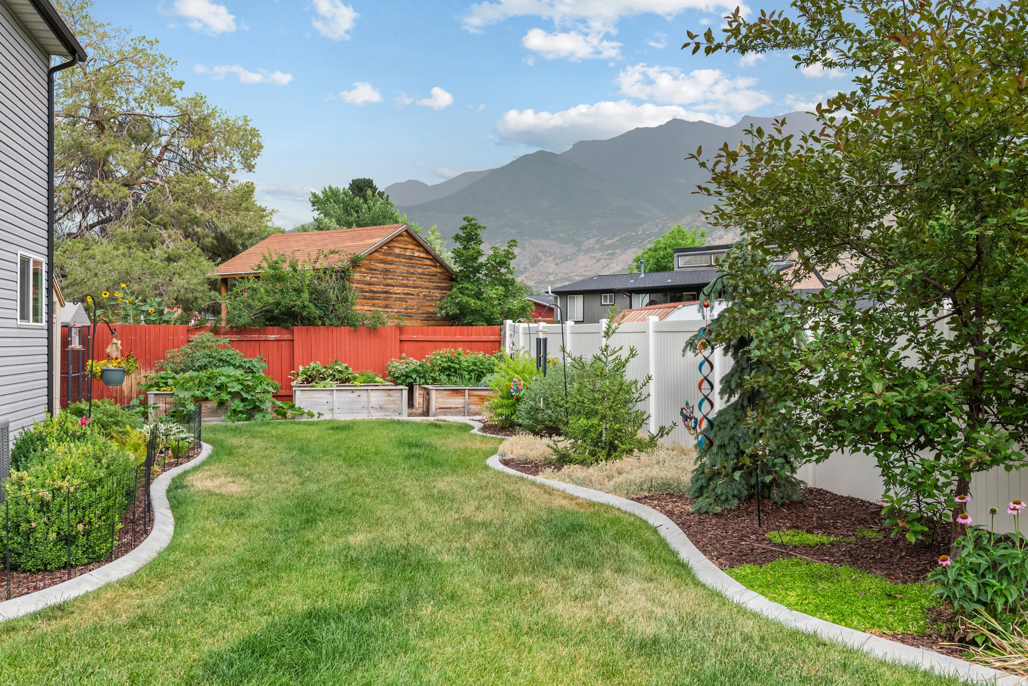 Expansive backyard featuring a manicured lawn, established landscaping, concrete curbing, a red privacy fence, and multiple raised garden beds