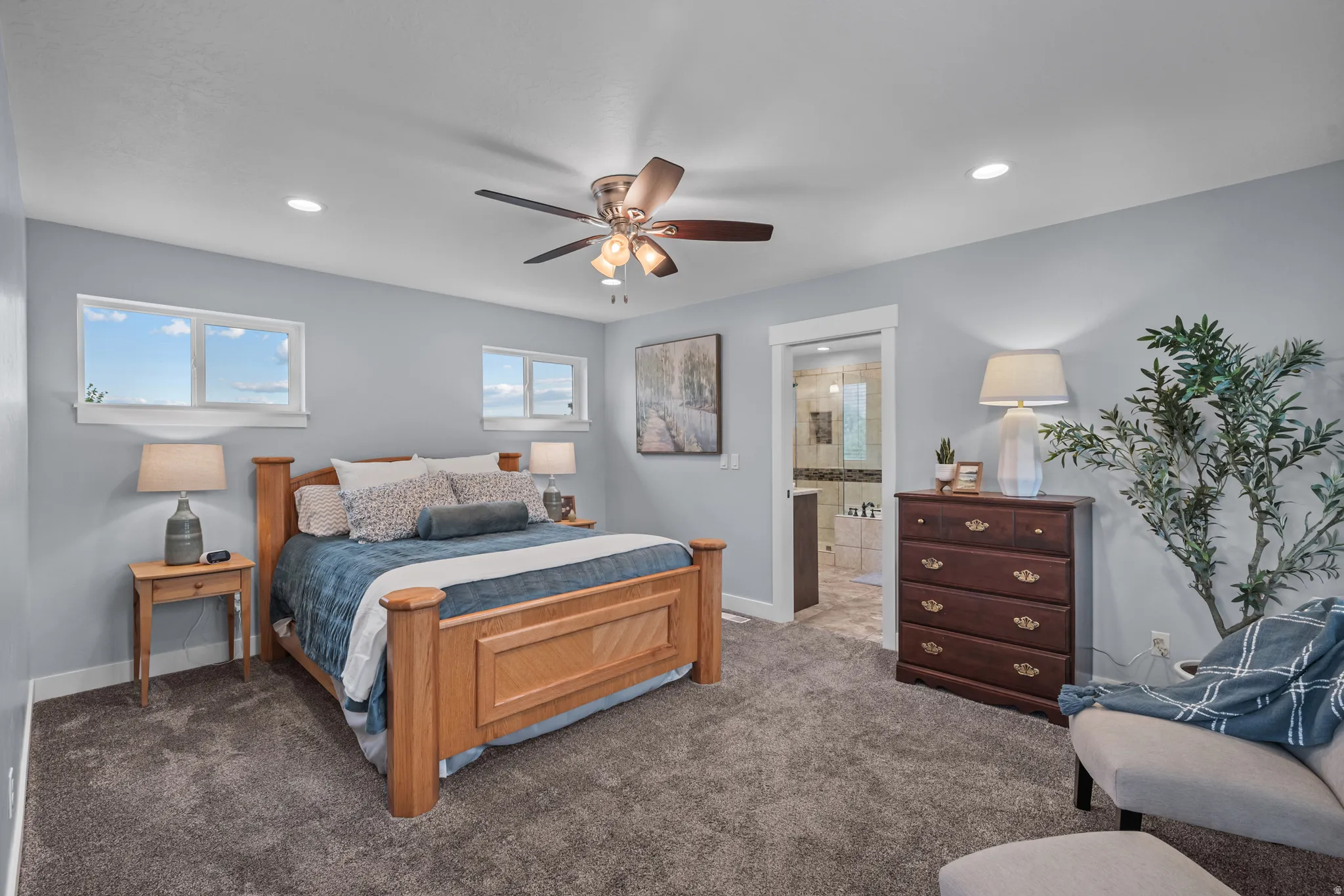 Carpeted bedroom featuring two windows with white trim, a ceiling fan with integrated lighting, recessed lighting, and a glass-enclosed shower visible through an open doorway