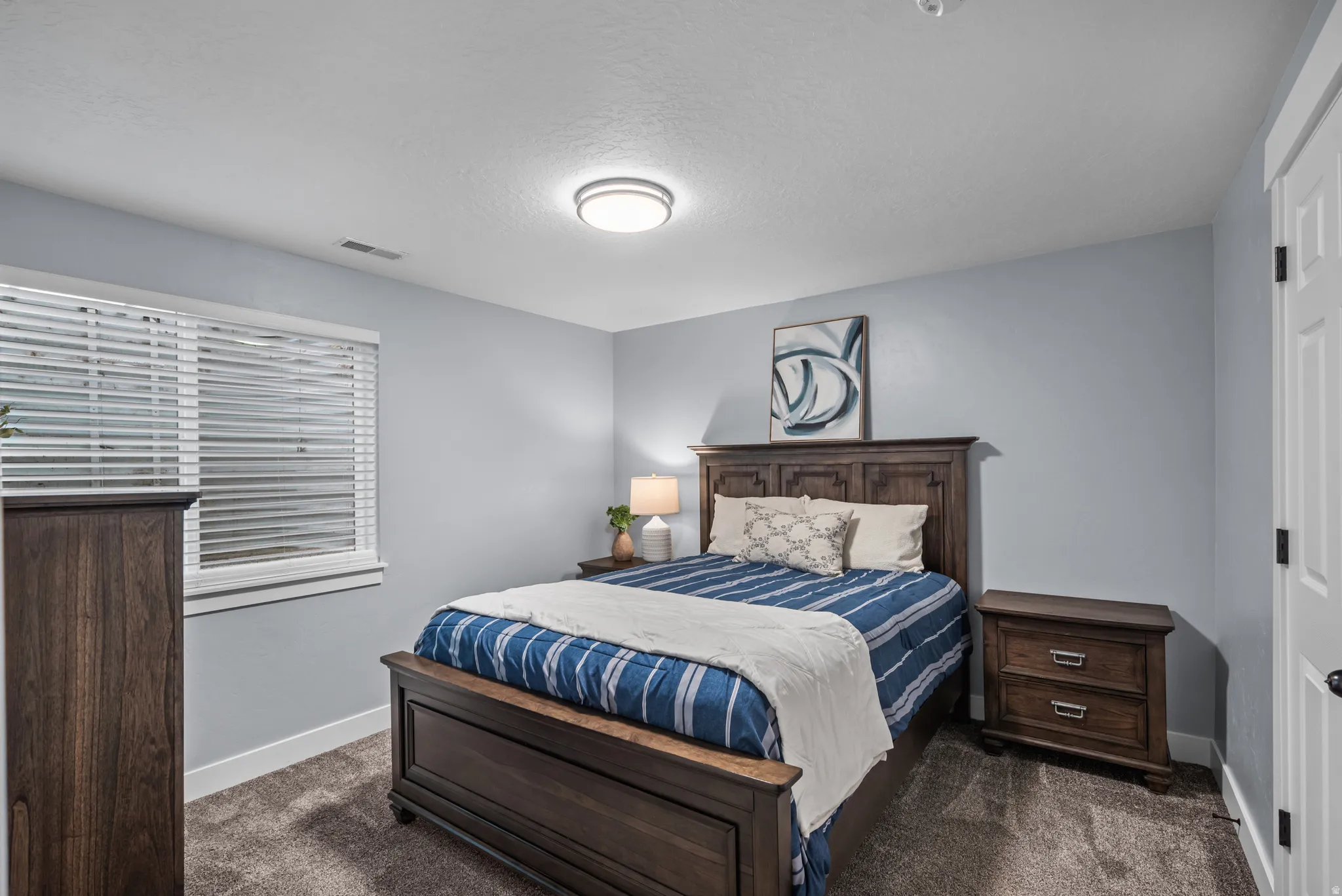 Carpeted bedroom featuring a window with horizontal blinds, light blue walls, and a ceiling-mounted light fixture