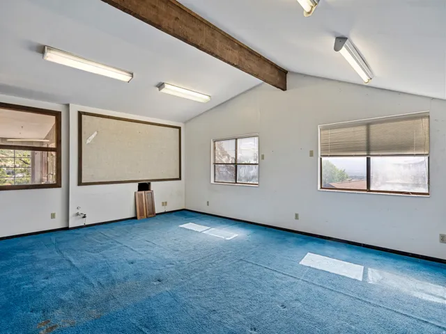 Spacious interior room featuring a vaulted ceiling with exposed wood beams and fluorescent lighting