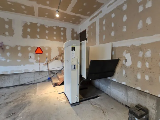 Utility room featuring a vertical platform lift, unpainted drywall, unfinished wood stairs, and a concrete floor