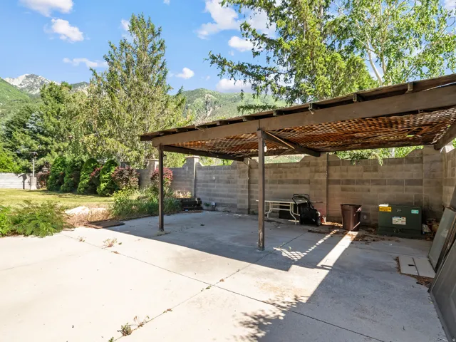 Concrete patio featuring a large pergola, surrounded by a block wall and mature trees