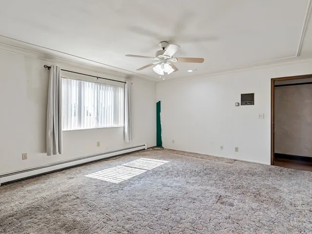 Spacious room with a ceiling fan, crown molding, baseboard heating, and a window with vertical blinds