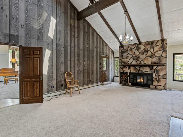 Spacious living area featuring a prominent stone fireplace with a wood mantle, vaulted ceilings with exposed wood beams, and vertical wood-paneled accent walls