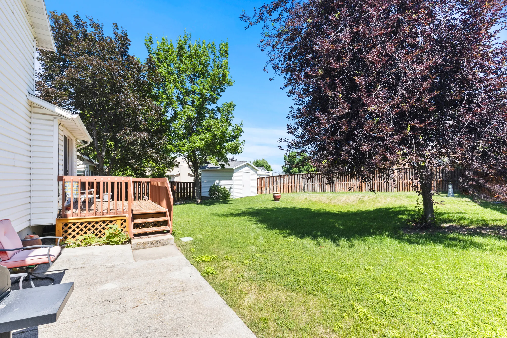 Expansive backyard featuring a wood deck with lattice detailing, concrete patio, mature trees, and a storage shed