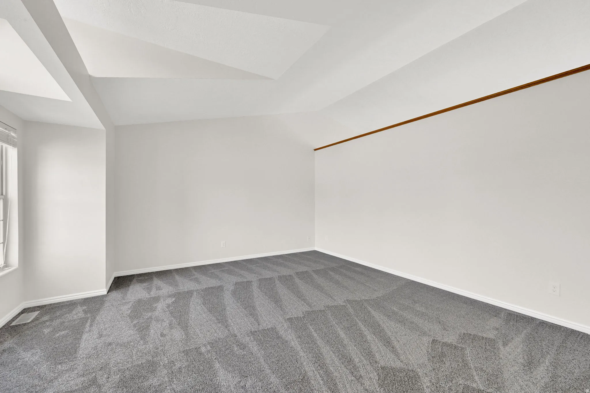 Spacious room featuring vaulted ceilings, a large window with white blinds, and patterned gray carpeting