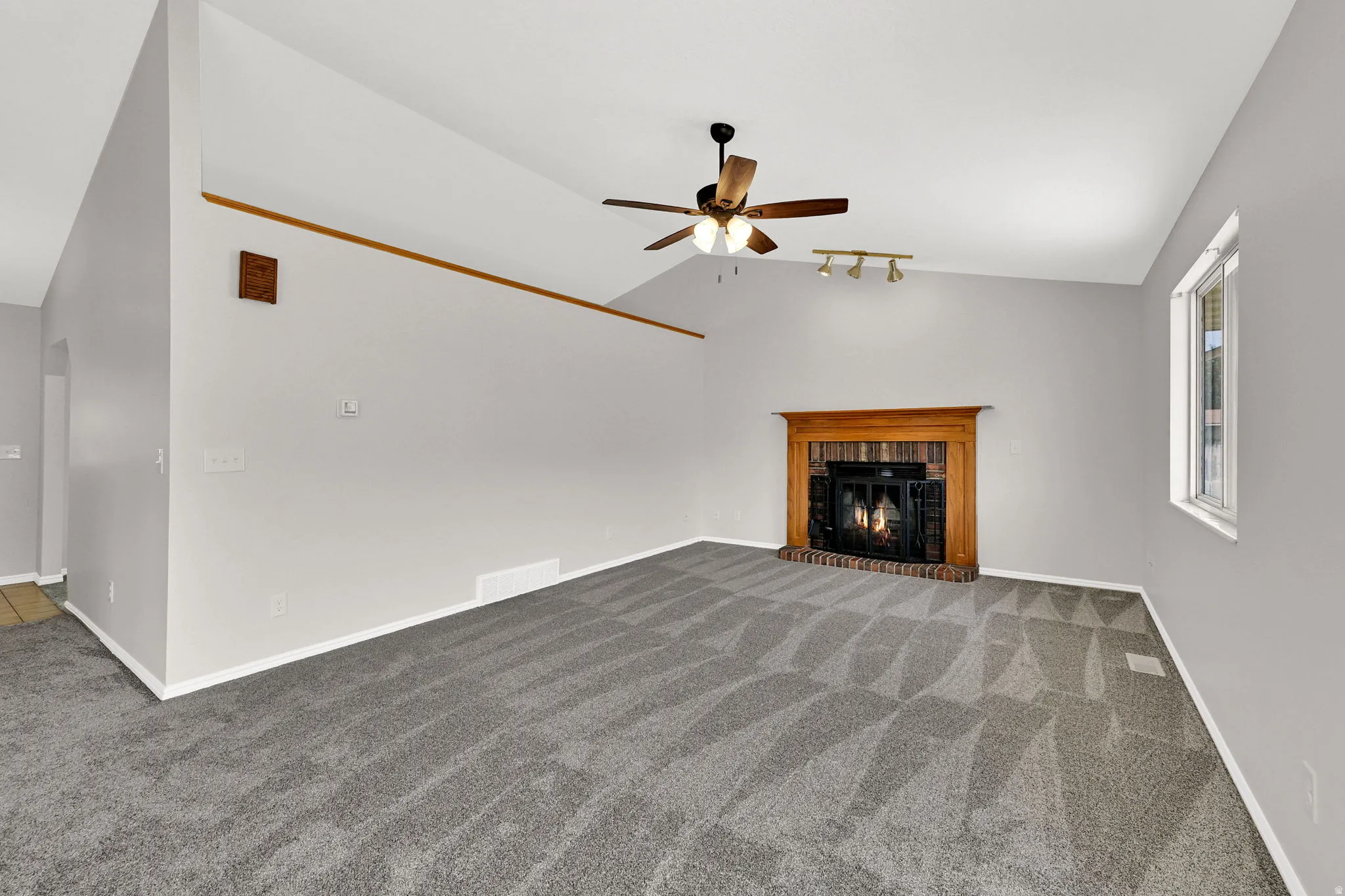 Spacious interior featuring vaulted ceilings, a wood-burning fireplace with a brick surround and wood mantel, a ceiling fan, and patterned grey carpet