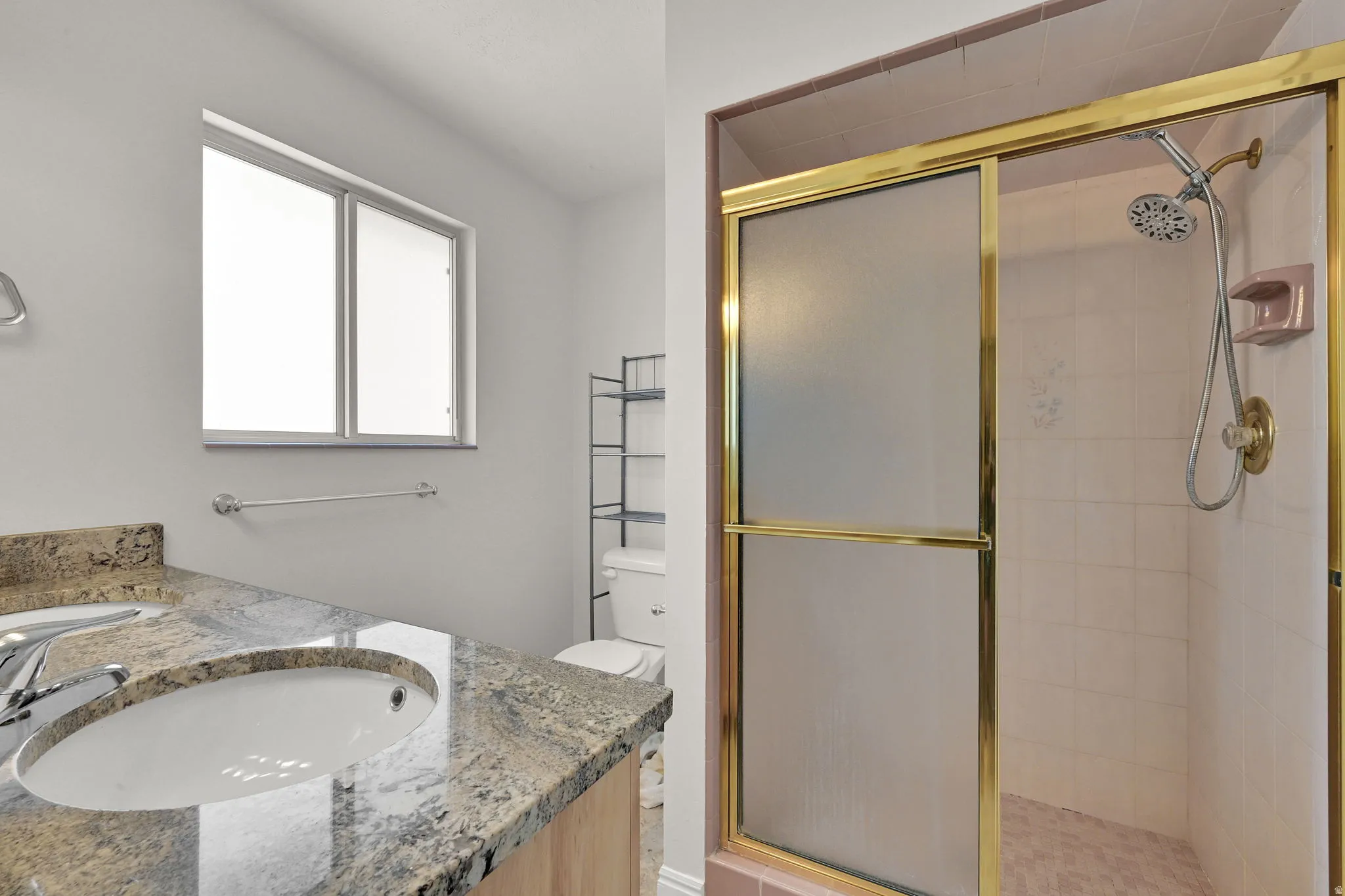Bathroom featuring a granite-finish vanity with an undermount sink, a large window, and a shower enclosure with a sliding frosted glass door and a gold-tone frame