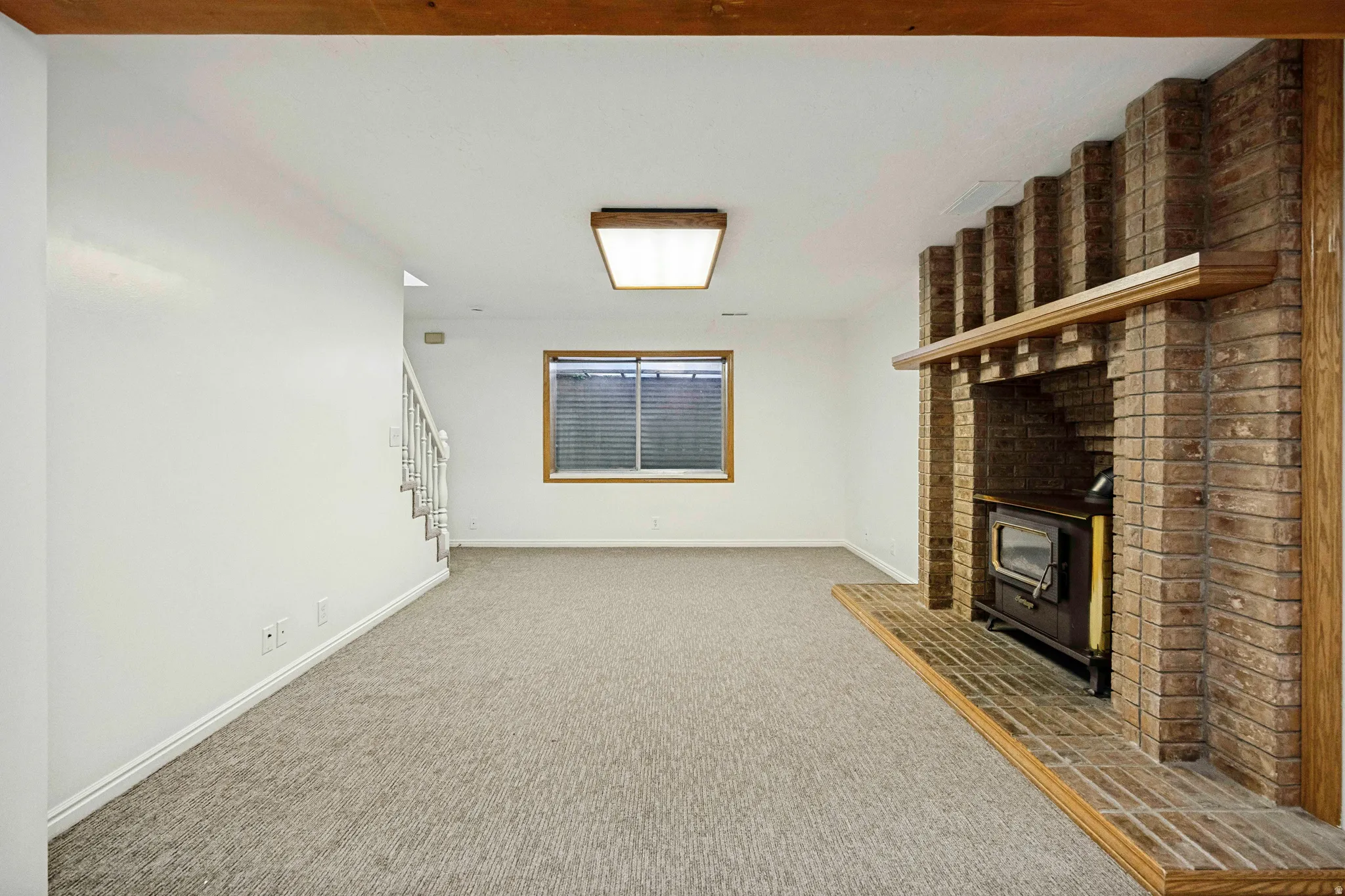 Spacious room featuring a brick fireplace with wood-burning stove insert, light-colored carpeting, white walls, and a window with blinds