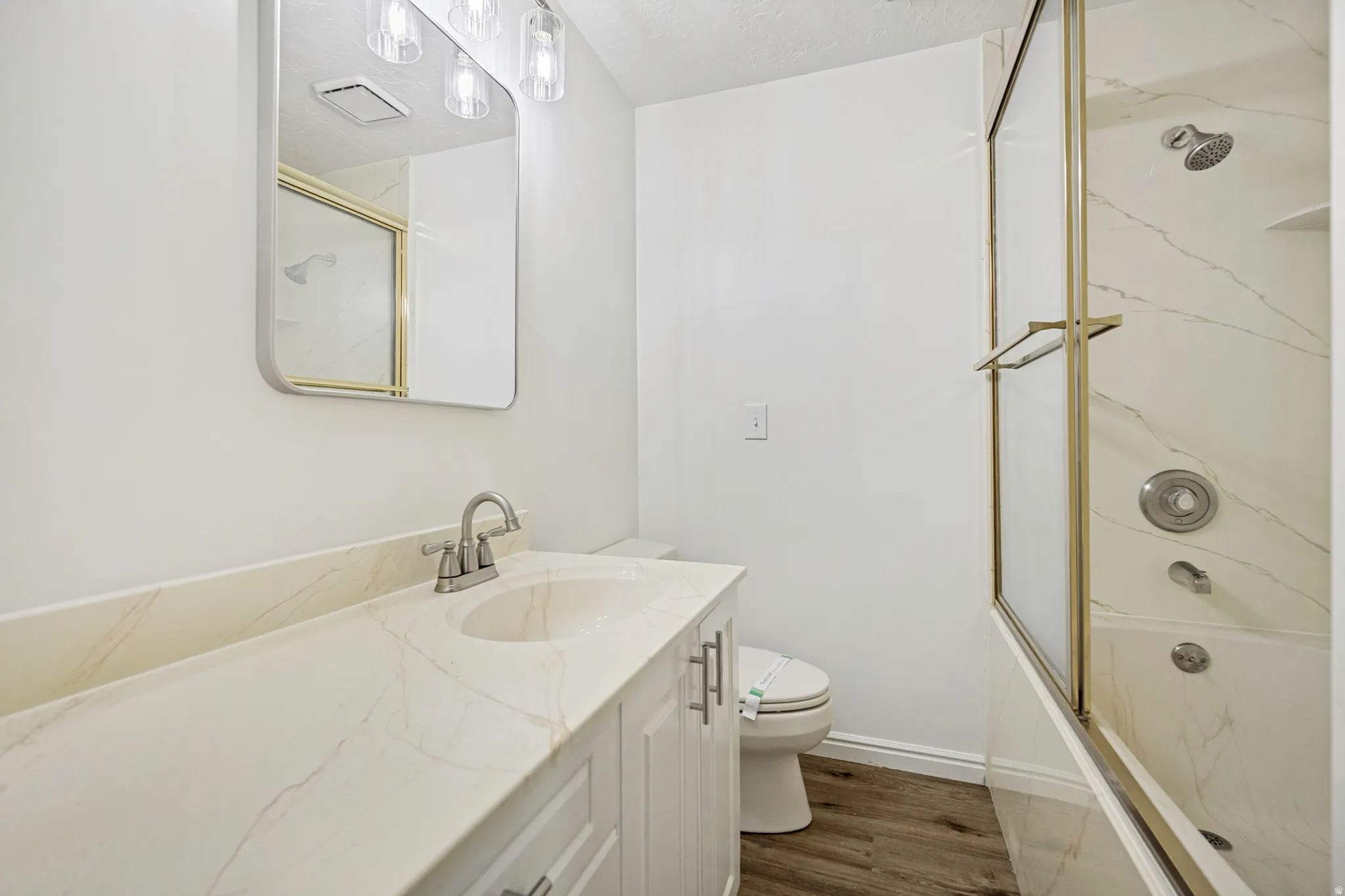 Bathroom featuring a white vanity with an integrated sink, a gold-framed shower/tub combination with a glass enclosure, wood-finish flooring, a rectangular mirror, and a three-light vanity fixture