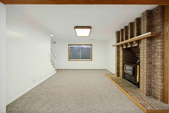 Spacious room featuring a brick fireplace with wood-burning stove insert, light-colored carpeting, white walls, and a window with blinds
