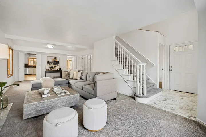 Spacious living area featuring light gray carpeting, white walls, a white staircase with balusters, and a front entry with a panel door and transom windows