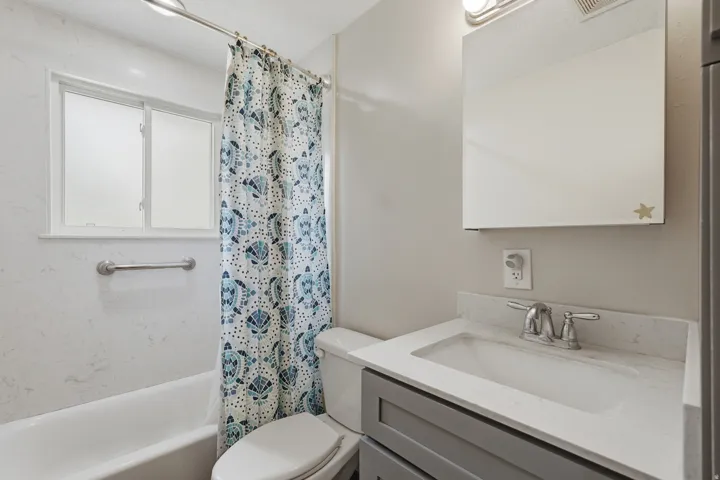 Contemporary bathroom featuring a white bathtub, frosted window, chrome grab bar, vanity with gray cabinetry, and a white countertop with an integrated sink