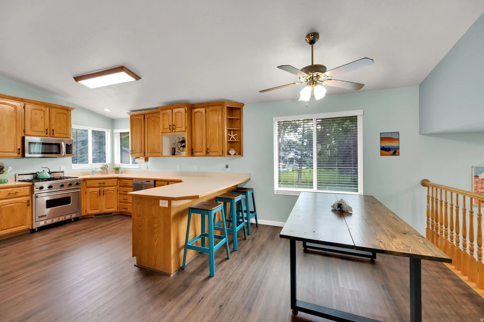 Spacious kitchen featuring wood-finish flooring, light wood cabinetry, a large kitchen island with seating, stainless steel appliances, and a window with blinds