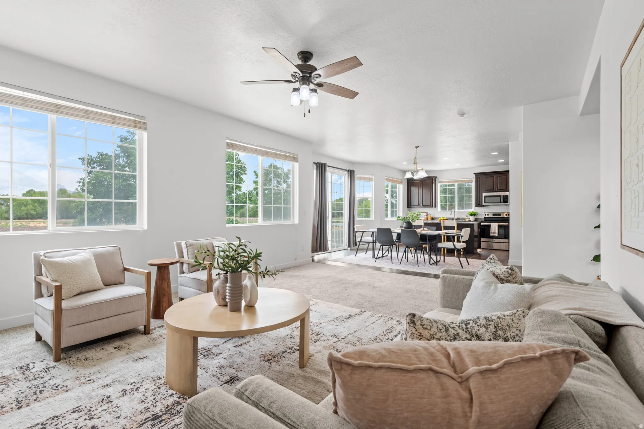 Light-filled living area featuring a ceiling fan with light fixture, large grid-pane windows, carpeted flooring, and an open concept layout to the kitchen and dining areas