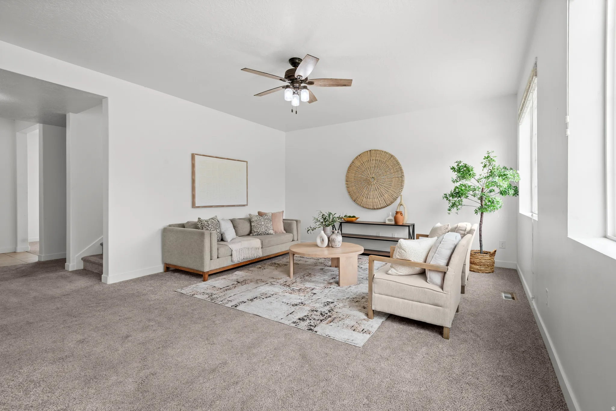 Spacious living area featuring neutral carpeting, white walls, multiple windows, and a ceiling fan with integrated lighting