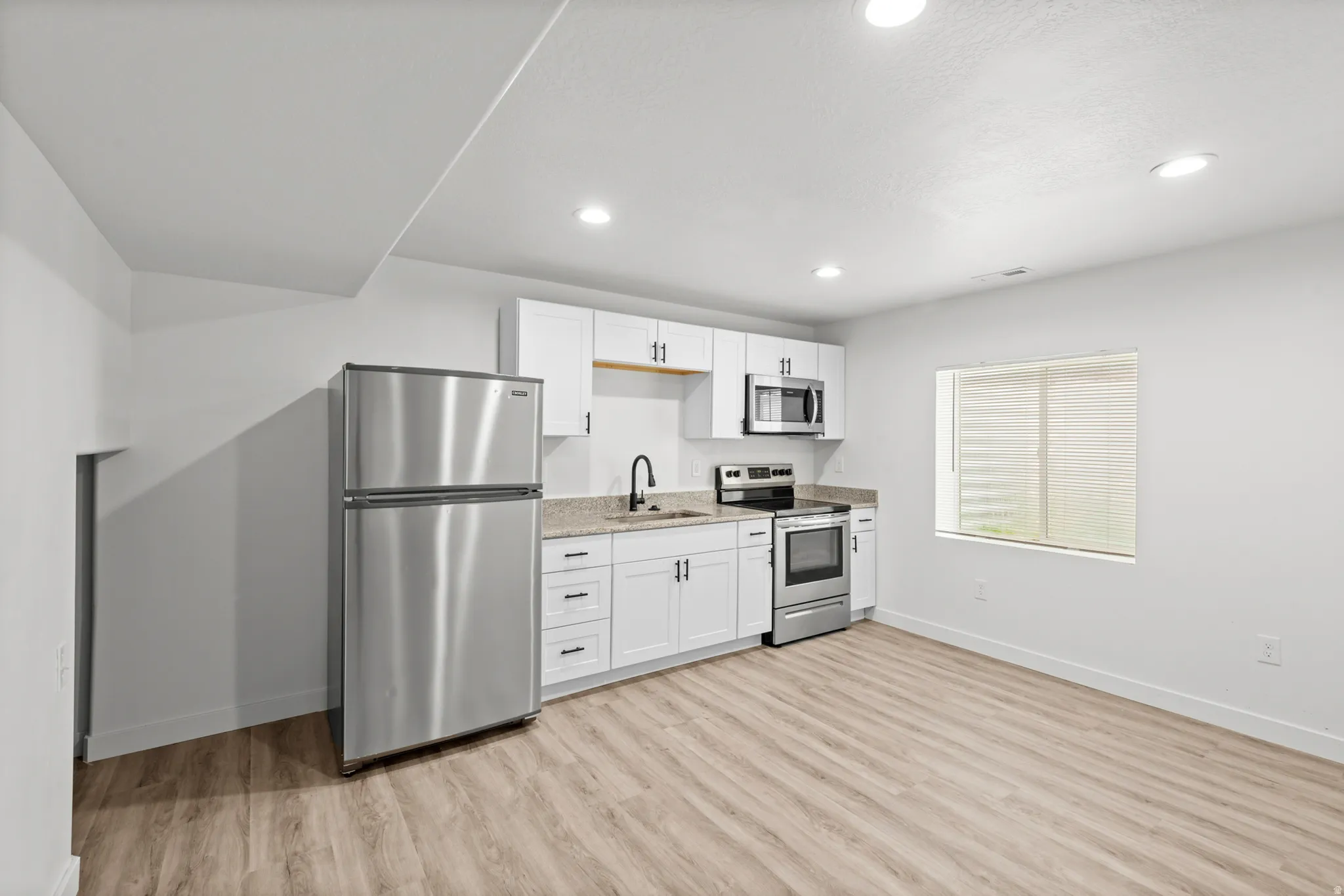 Contemporary kitchen area featuring white cabinetry, stainless steel appliances, granite-style countertops, wood-finish flooring, and recessed lighting
