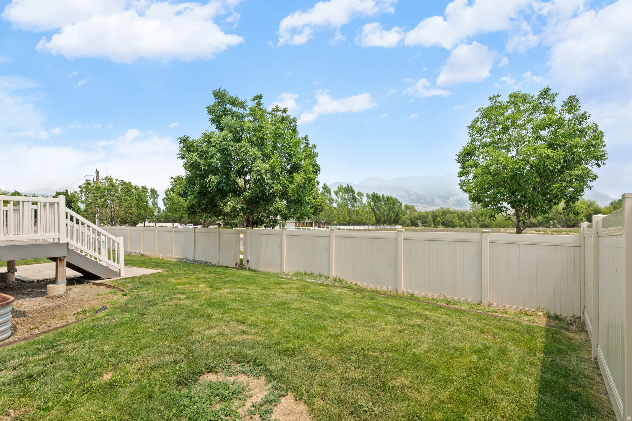 Expansive backyard featuring a large green lawn, a multi-level deck with white railings, a tan privacy fence, and mature trees