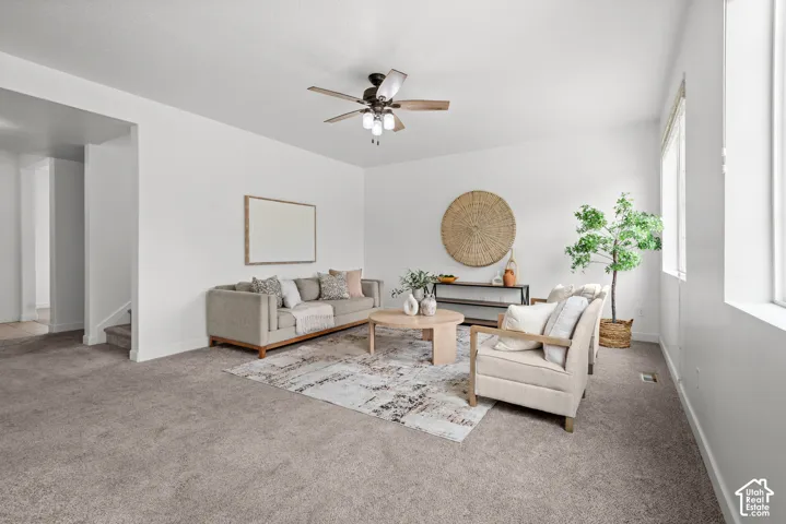 Spacious living area featuring neutral carpeting, white walls, multiple windows, and a ceiling fan with integrated lighting