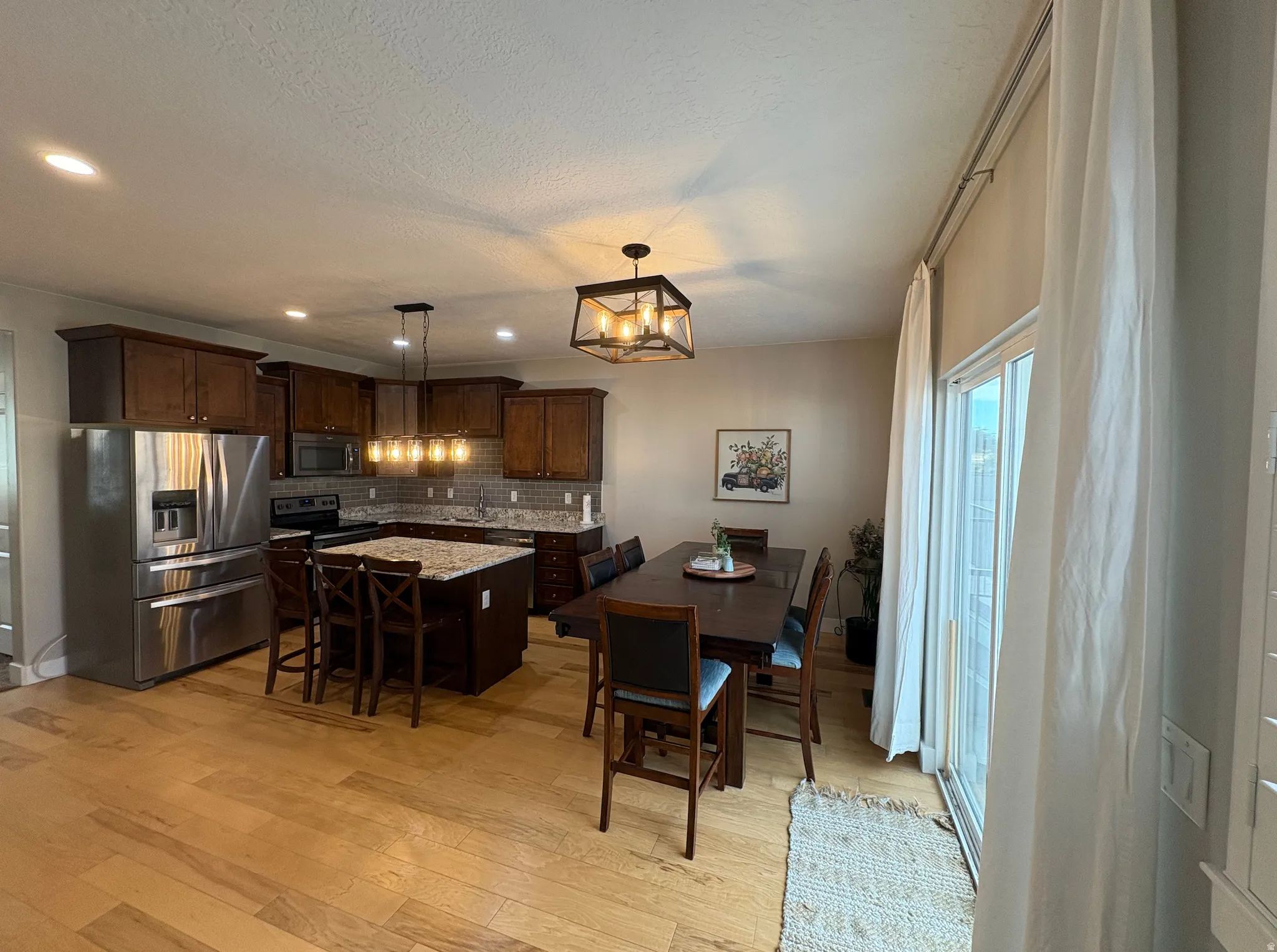 Kitchen and dining area featuring wood-finish flooring, dark wood cabinetry, a central island with a light-toned countertop, stainless steel appliances, and a sliding glass door