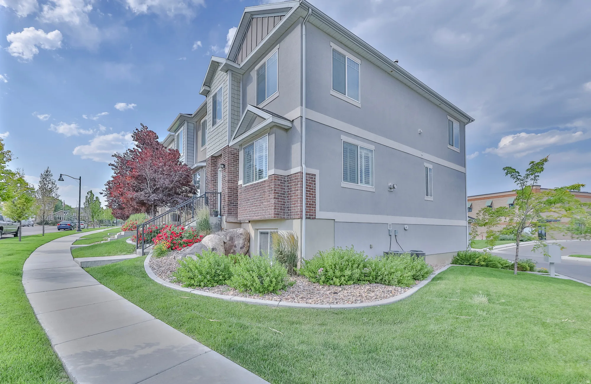 Multi-story townhouse exterior with light gray stucco and siding, brick accents, white frame windows, and a landscaped front yard featuring mature shrubs and decorative rocks