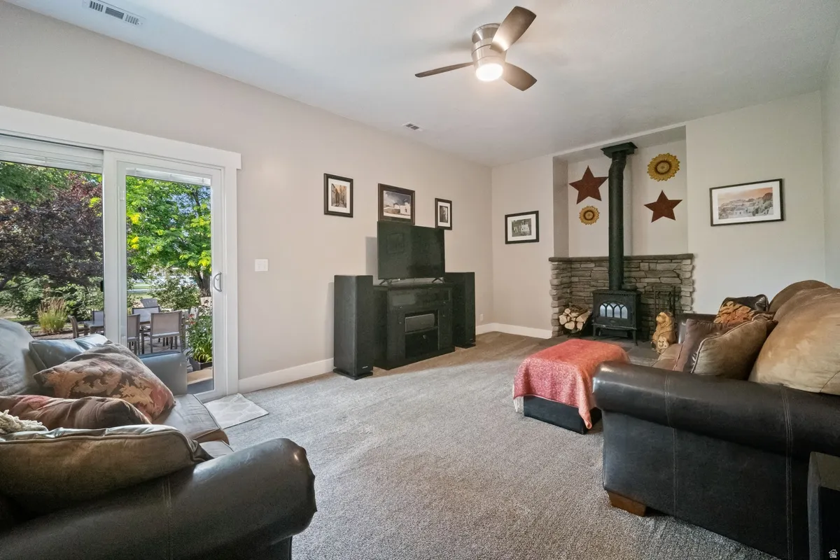 Carpeted living area featuring a stone accent wall with a wood-burning stove, a ceiling fan with integrated lighting, and a sliding glass door leading to an outdoor patio with surrounding greenery