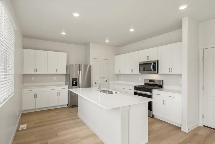Modern kitchen featuring a central island with an undermount sink, white shaker cabinetry, stainless steel appliances, and light wood-finish flooring
