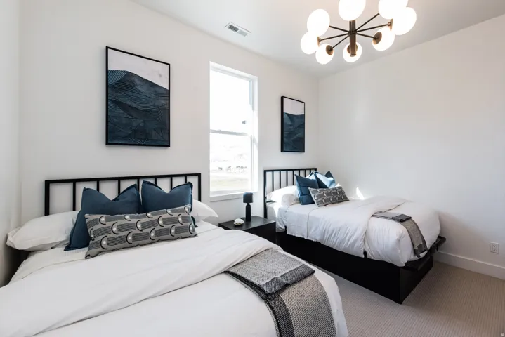 Bright room featuring two beds with black metal headboards, a large window, a modern chandelier, and light-toned carpet flooring
