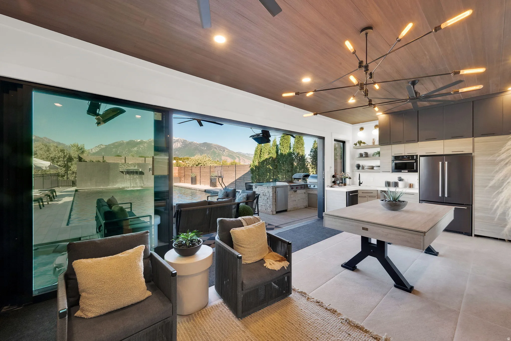 Expansive indoor-outdoor living space featuring floor-to-ceiling sliding glass doors, a wood-finish ceiling, and a modern linear chandelier