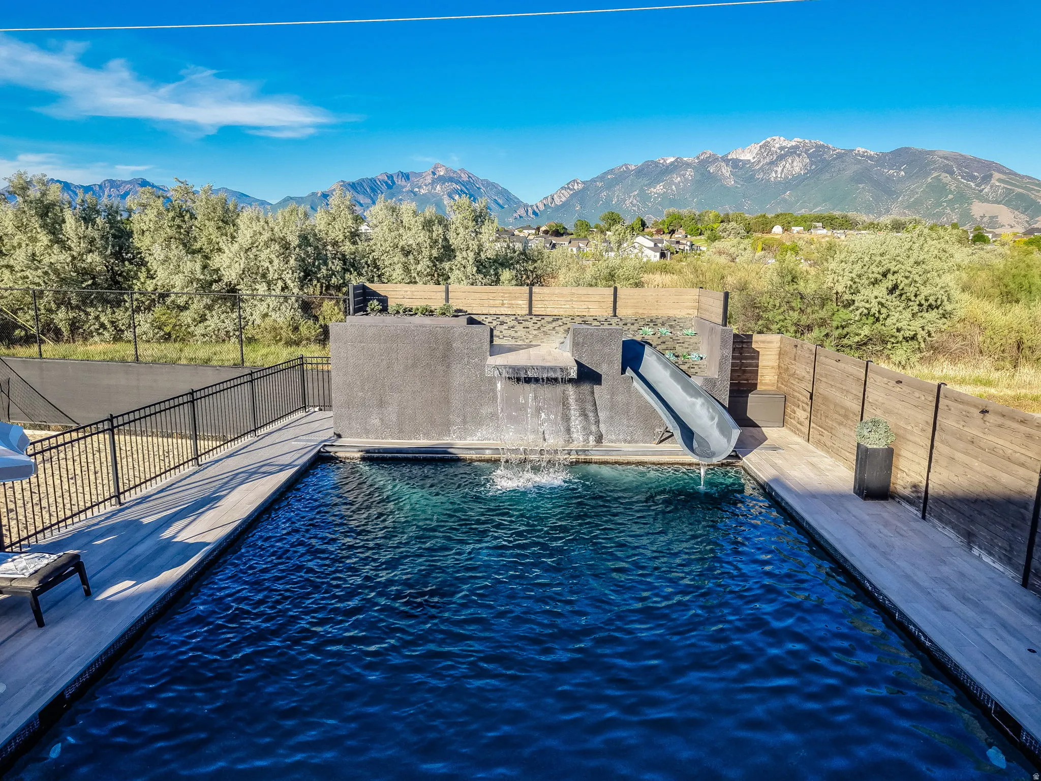 Outdoor swimming pool with a dark finish, integrated slide, waterfall feature, and surrounding wood-finish decking