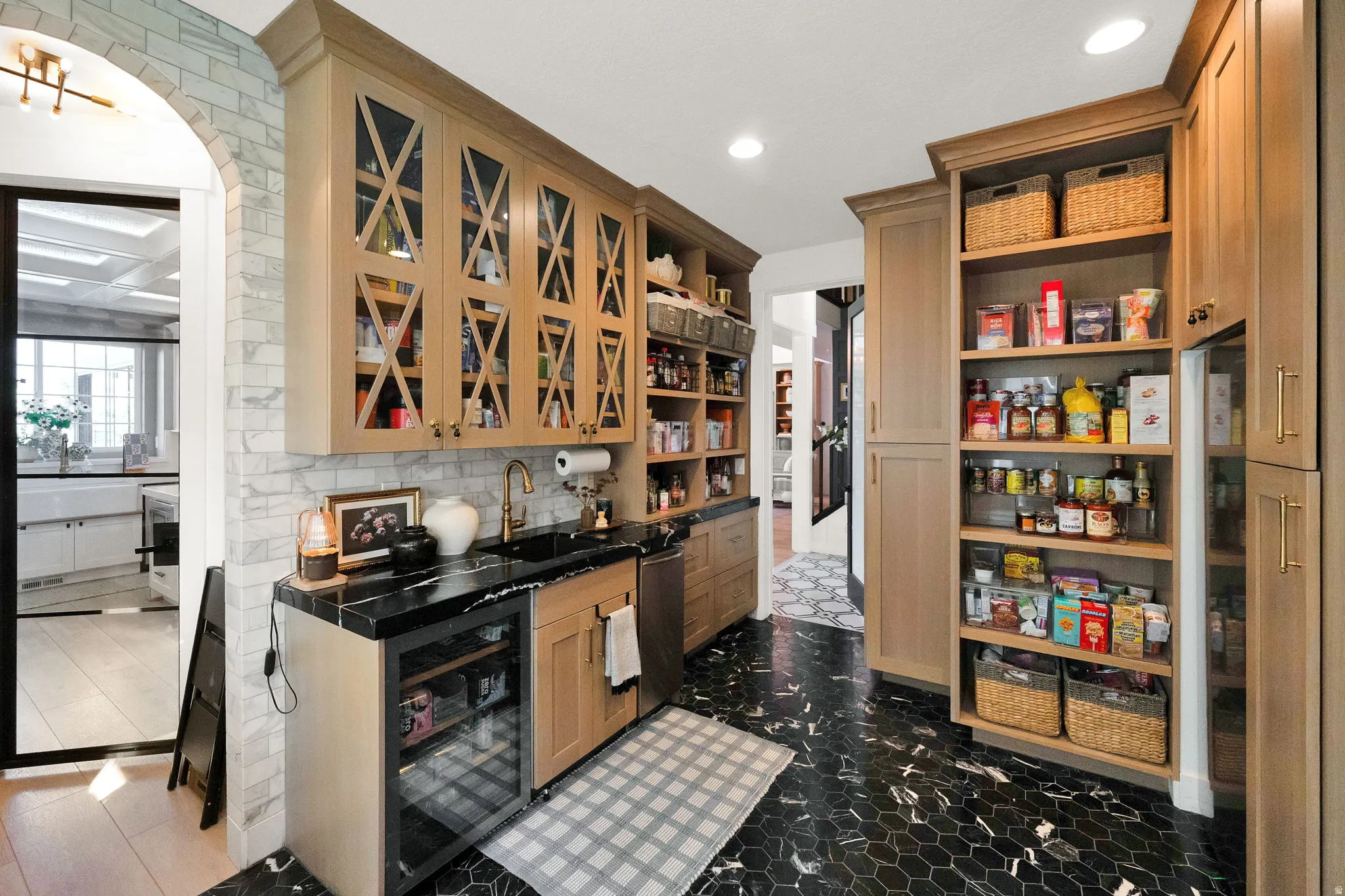 Gourmet pantry featuring custom wood cabinetry, integrated wine cooler, dark stone countertops, undermount sink with brass-finish faucet, and black hexagonal floor tiling