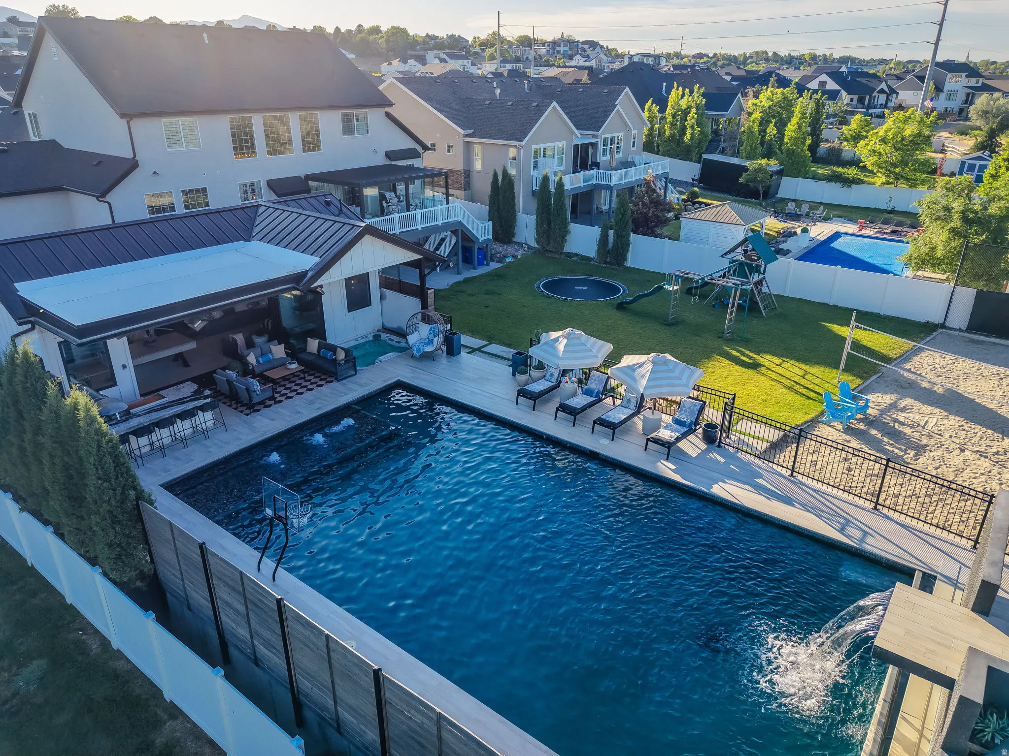 Expansive backyard featuring a rectangular swimming pool with integrated fountains and a waterfall, a covered outdoor living area with a bar, a sand volleyball court, and a manicured lawn