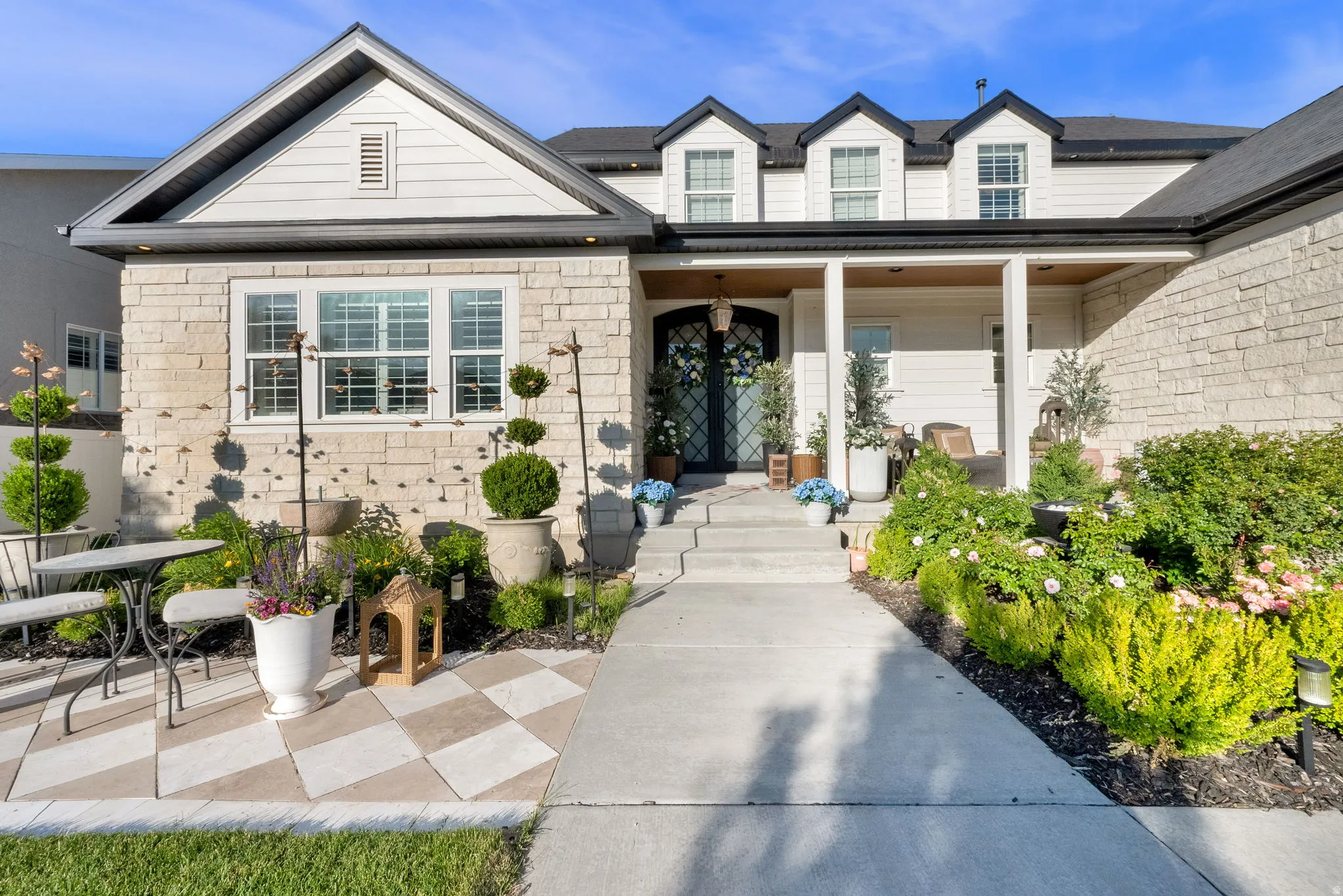 Stone and siding exterior featuring a decorative front door, covered porch, multiple dormer windows, and a checkered patio area